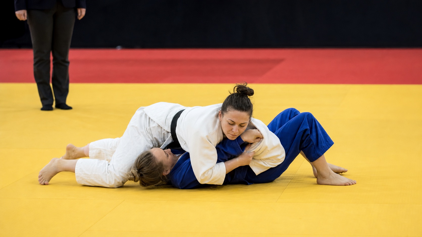 Two female judoka contesting a ne-waza hold-down on the tatami.